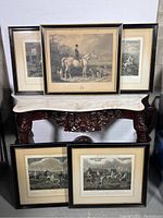 Photo showing five framed antique equestrian etchings arranged on and in front of a marble-topped wooden table, frames are black with gold trim, images are sepia toned with riders, horses and dogs.