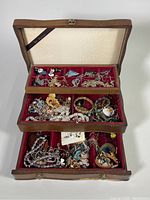 Wooden jewelry box opened showing three sections filled with assorted costume jewelry including necklaces, bracelets, bangles, earrings, and pins.