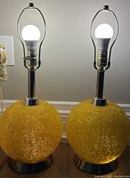 Pair of Mid-Century Modern table lamps with glowing bulbs on, showing illumination and lamp structure with yellow lucite orb bases and brass-coloured metal supports.