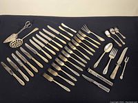 Photo showing entire collection of cutlery and serving utensils laid out on black surface, including knives, forks, small spoons, slotted spoon, and cake tongs.