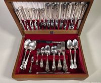View of entire silver plate flatware set inside wooden chest with red velvet lining and satin fabric insert on lid. Shows rows of knives, forks, spoons, and serving spoons.