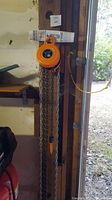 Full view of orange chain hoist hanging from wooden support showing chains and hook.