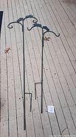 Two tall metal double shepherd's hooks laid on outdoor wooden deck, showing full length and stakes for ground insertion.