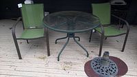 Two green sling fabric patio chairs with metal armrests and legs, a round glass top metal table with a 40 inch diameter, all set on a wooden deck.
