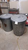 Two galvanized metal garbage cans viewed from front, showing lids and handles. One can has a rust patch visible on the body.