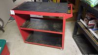 Full view of the red metal rolling work bench with rubber mat on top and two shelves below.