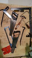 Photo of assorted hand tools including two handsaws, a claw hammer, coping saw, screwdriver, rasp file, try square, and clamp arranged on a wooden surface with a black measuring square.