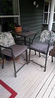 Photo of two metal frame bar height chairs with floral and grey cushions around a bar height square table on a wooden porch decking. The table holds a small radio and a potted plant.