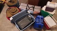 Photo showing two rolled sleeping bags, blue plastic bin with air mattress, three coolers, large blue water jug, and tent bag.