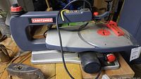 Front and side view of Craftsman scroll jigsaw on wooden surface, showing the entire tool including power cord and motor casing, red speed control knob, black and grey body.
