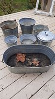 Photo of six metal pails and one metal tub with dirt and leaves inside, arranged on a wooden deck, varying in size.