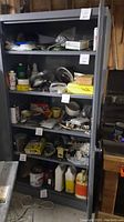 Front view of the metal storage cabinet showing five shelves filled with various miscellaneous items such as bottles, tools, tapes, and containers. Shelves are metal and loaded with mixed objects, not included in sale.