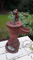 Front angle view of red painted cast iron water pump showing handle, spout, and base with rust and wear.