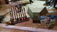 Full view of croquet set with mallets, balls, metal stand, wood totem pole with blue wings, metal firewood holder, and rolled shade on floor