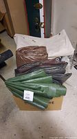 Several tarps folded and stacked in a box, showing green, brown, gray, and white tarps with grommets visible.
