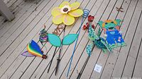 Overview of all six whimsical yard art items on deck: rainbow hot air balloon, yellow flower, blue spiral stake, red hummingbird feeder, sea turtle, frog with overalls.