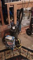 Photo of the wet/dry shop vacuum, Hartz spotlight box, and work light with clamp on floor under wooden frame in workshop setting.