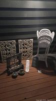 Photo showing two ornate metal wall panels, a white wicker chair, a decorative fireplace grate, two small pots, a white garden thermometer or sign, and a black decorative garden plaque.