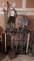 Overview of assorted yard tools stored vertically on black rack with white legs against the wall, including shovels, rakes, hoes, and a plastic scoop.