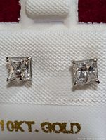Close-up photo of pair of 10K gold stud earrings with square-cut clear cubic zirconia stones set in four-prong settings, shown on white textured jewelry holder with '10KT GOLD' marking visible.
