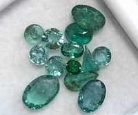 Close-up of 12 loose emerald and aquamarine gemstones showing varied oval and round cuts and shades of green and blue.