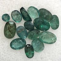 Fifteen loose emerald gemstones laid out on a white textured background showing variety of shapes and inclusions