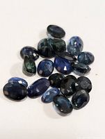A collection of eighteen loose faceted genuine sapphire gemstones in various shapes and sizes, showing deep blue to greenish-blue hues on a white background.