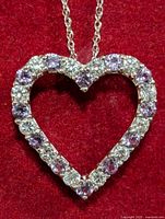 Close-up of silver heart-shaped pendant on red background showing alternating simulated alexandrite (purple) and clear stones.