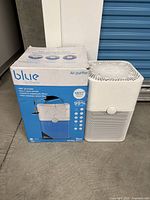White Blueair Pure 211+ air purifier next to its original blue and white box with some visible damage.