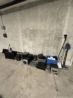 Full view of assorted electronics lot including speakers, audio mixer, vacuum cleaners, and bins of accessories lined up against a concrete wall.