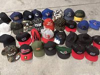 Photo showing the full arrangement of 26 assorted caps in three rows on concrete floor, various styles, colors, and logos visible.