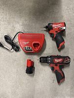 Overview of all items: M12 charger, battery pack, cordless drill/driver, and FUEL impact driver on concrete floor