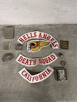 Top-down photo showing all memorabilia including three Hells Angels patches, two rectangular patches, and various metal accessories arranged on a concrete floor.