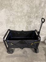Photo of a black collapsible moving cart with fabric basket, four wheels, and telescopic handle against a gray concrete wall.