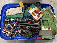 Top view of blue bin containing assorted vintage LEGO pieces and baseplates including green and grey platforms, red and yellow brick pieces, and black ramp elements.