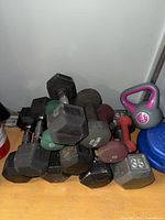 Photo of assorted dumbbells stacked on floor including black hex dumbbells (10 lb, 35 lb), green, maroon, and orange dumbbells.
