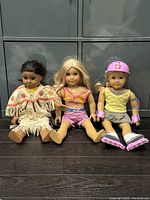 Three American Girl dolls sitting on black floor in front of a gray cabinet, showing full body front view, dressed in distinct outfits.