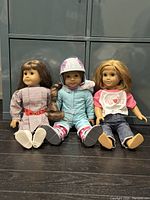 Front view of all three American Girl dolls sitting on the floor, showcasing their full outfits and details.