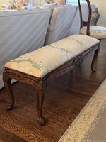 Side and partial top view of the antique upholstered bench, showing curved cabriole legs, ornate carvings, and floral patterned upholstery in pastel colors with nailhead trim.