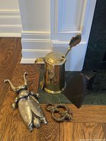 Photo showing the brass firestarter pitcher with lid and wand, solid brass beetle boot jack, and iron doorstop on wood floor.