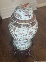 Full view of the large ceramic urn with lid and wooden stand, showing the overall shape and decoration including the butterfly handle