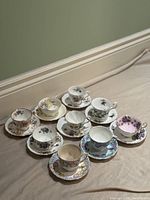 Nine fine china tea cups with matching saucers displayed on a beige fabric surface near a green wall.