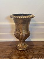 Front view of small gold urn with black interior sitting on wooden floor against white baseboard and wall.