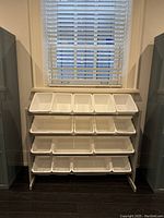 Front view of a grey rack with 21 white angled bins of varying sizes arranged in three rows beneath a window.