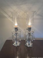 Pair of vintage boudoir table lamps illuminated, showing etched glass hurricane shades and hanging crystal prisms on clear bases.