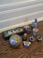 Photo showing all 8 vintage enamel and collectible pill boxes displayed on wood floor including rectangular painted village scene box, cloisonné round and butterfly-shaped boxes, and others