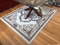 Photo showing entire rectangular area rug under glass table on wood floor, exhibiting beige and green patterned border with floral and geometric elements.