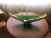 Top-side view of the green elongated art glass bowl on a wooden table near a window with sheer curtains.