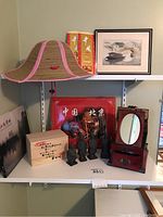 Shelf with red wooden jewelry box with mirror and drawer, small dark statues, red painted wooden box with Chinese characters, souvenir print of Asian boat scene, two books, woven sun hat with pink trim on top shelf