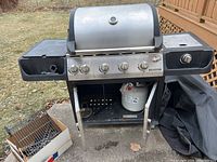 Full view of the Galveston 5-burner gas barbecue grill with side burner, propane tank under the grill, and a cardboard box containing extra accessories on the side shelf.